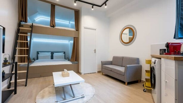 A compact hotel room with a bunk bed, a sofa, a small table, a round mirror, and a kitchenette. - Home Voltaire (Paris)
