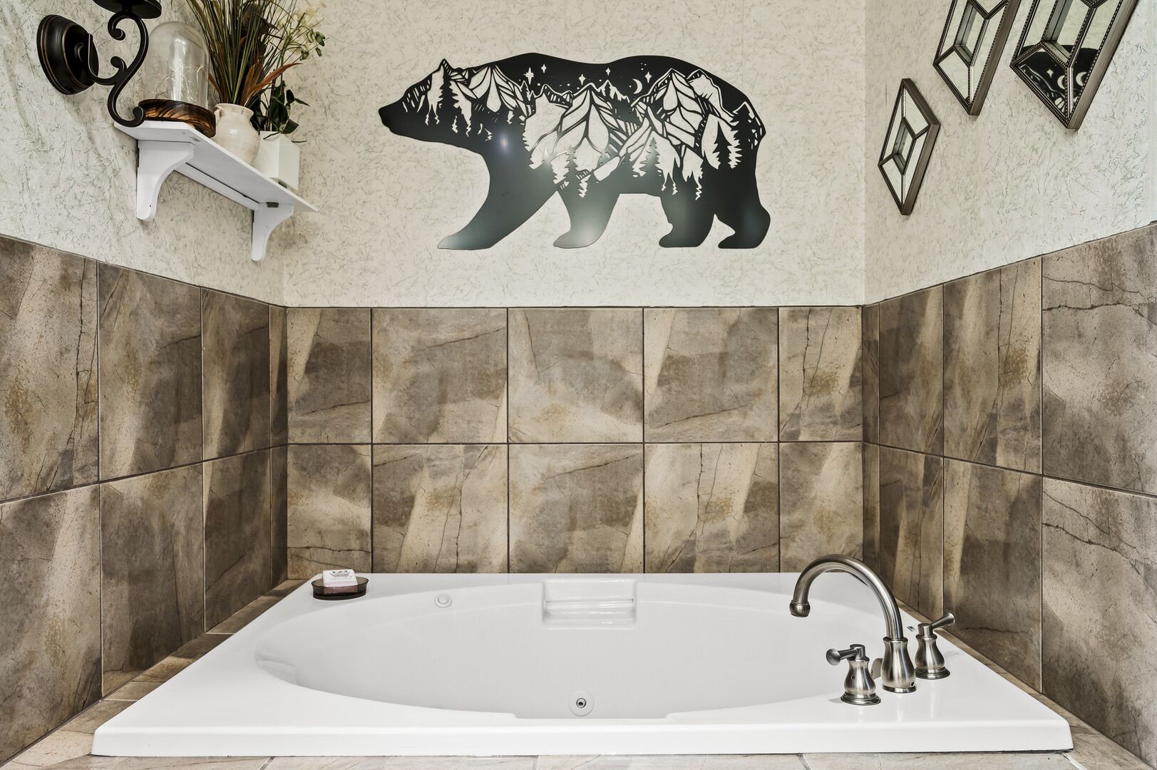 Un baño con una bañera grande, paredes de mármol, un mural de un oso y un estante con objetos decorativos.