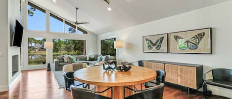 A spacious living area with a round wooden table and chairs, a sofa, a TV mounted on the wall, and two butterfly paintings.