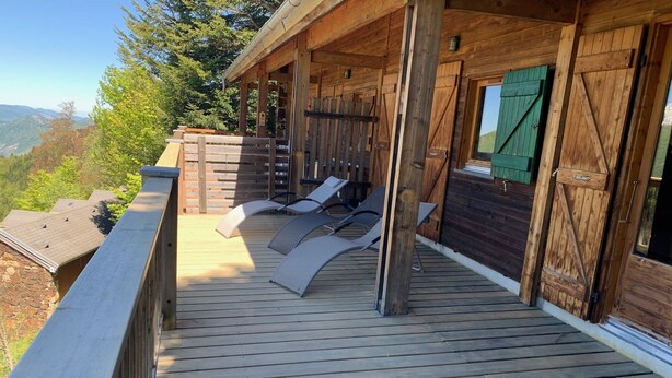 A wooden deck with lounge chairs, a wooden cabin with a green shutter, and a view of the surrounding landscape.