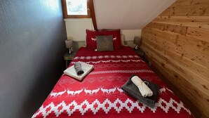 A cozy bedroom with a sloped ceiling, a wooden wall, a red bedspread with reindeer patterns, a nightstand with a lamp, and a window letting in natural light.