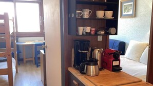 Cocina compacta con encimera de madera, cafetera y hervidor. Se ve un comedor con mesa y sillas a través de un puerta abierta.