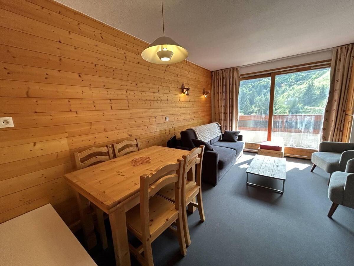 A cozy room with a wooden wall, a dining table with chairs, a sofa, a coffee table, and a large window with curtains.