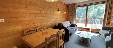 A cozy room with a wooden wall, a dining table with chairs, a sofa, a coffee table, and a large window with curtains.