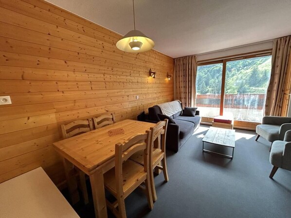 Une pièce chaleureuse avec un mur en bois, une table à manger avec des chaises, un canapé, une table basse et une grande fenêtre avec des rideaux.