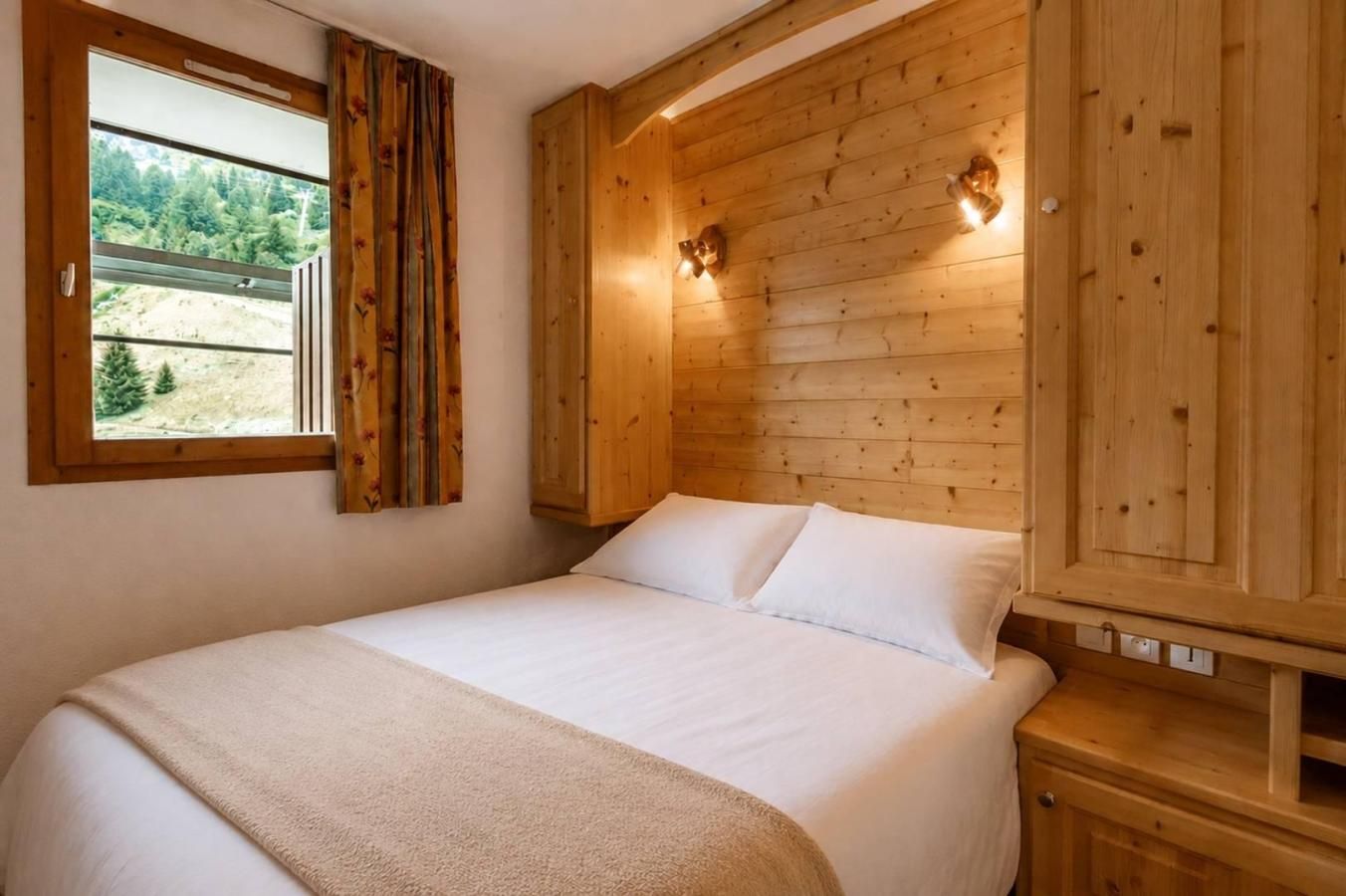 A bedroom with a wooden headboard, a bed with white linens, a window with curtains, and a view of trees and a hill.