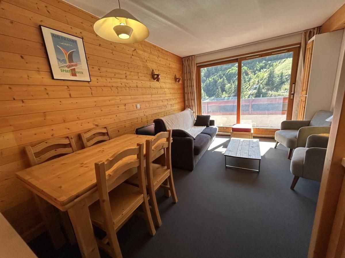 A room with a wooden dining table, chairs, a sofa, a coffee table, and a large window with a view of greenery.