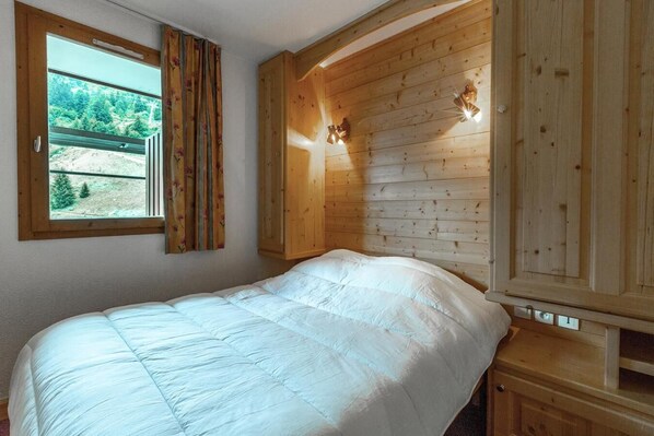 A bedroom with a wooden headboard, a large window with curtains, and a bed with a white duvet.