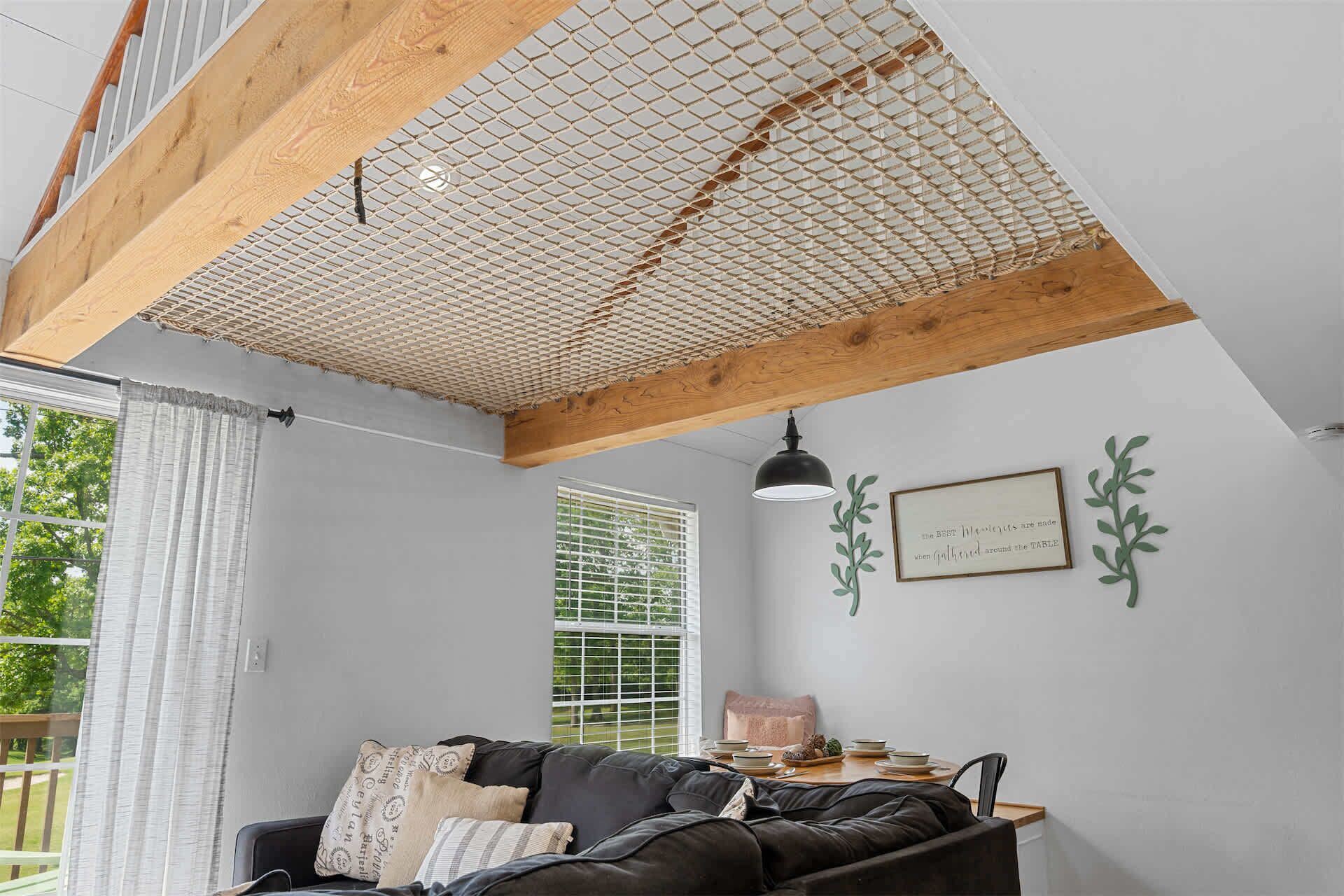 A room with a black leather sofa, a dining table with dishes, a window with blinds, and a ceiling with a wooden beam and mesh.