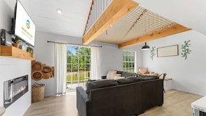 Een moderne woonkamer met een donkere bank, houten balken, een open haard en uitzicht op een groen buitengebied.