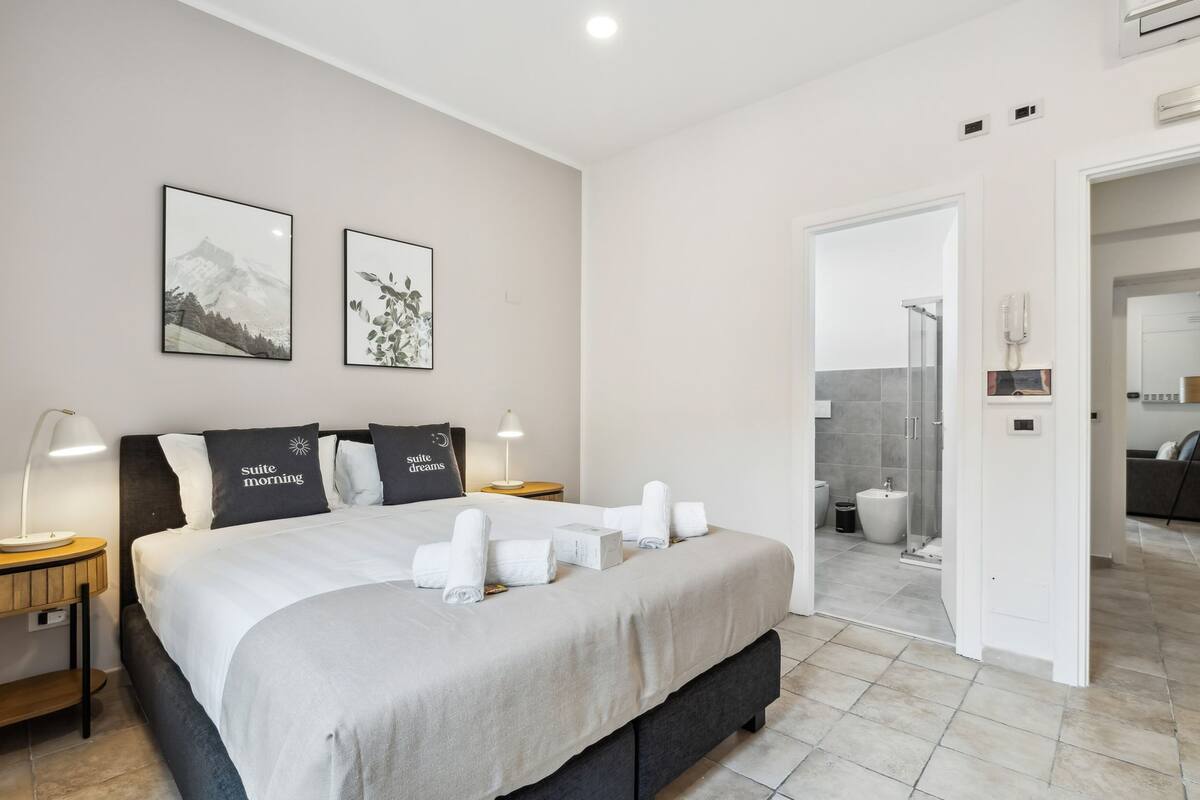A modern hotel room with a large bed, two bedside tables with lamps, a shower area, and a bathroom with a toilet and sink.
