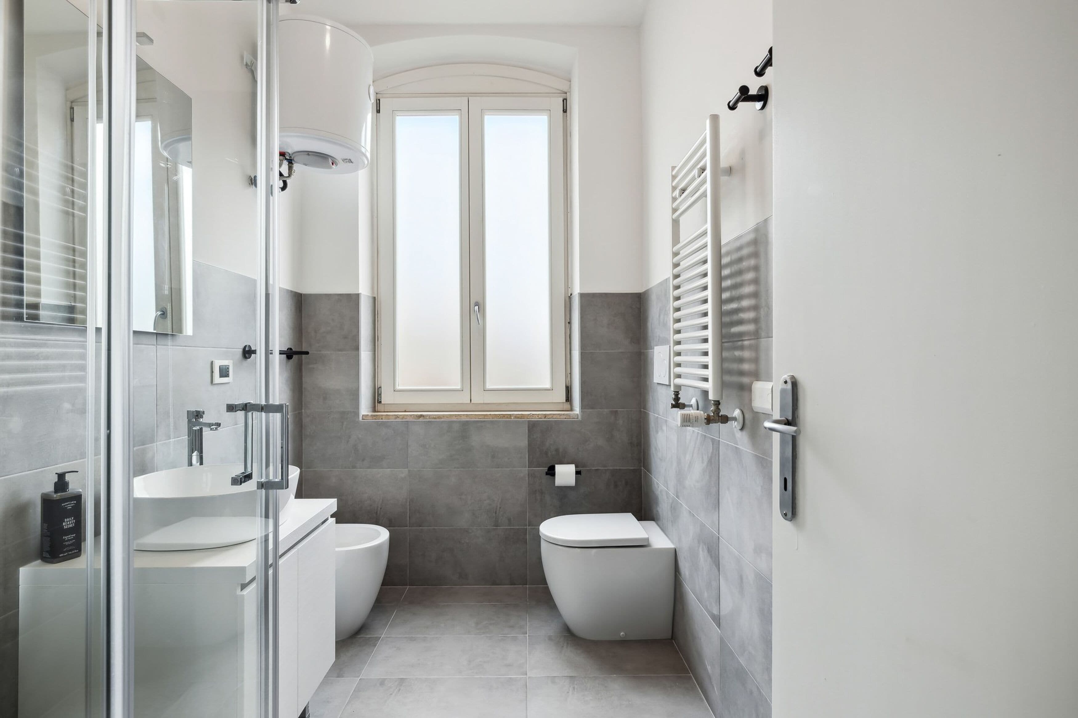 A modern bathroom with a glass shower enclosure, a white sink, a toilet, and a window above the toilet.