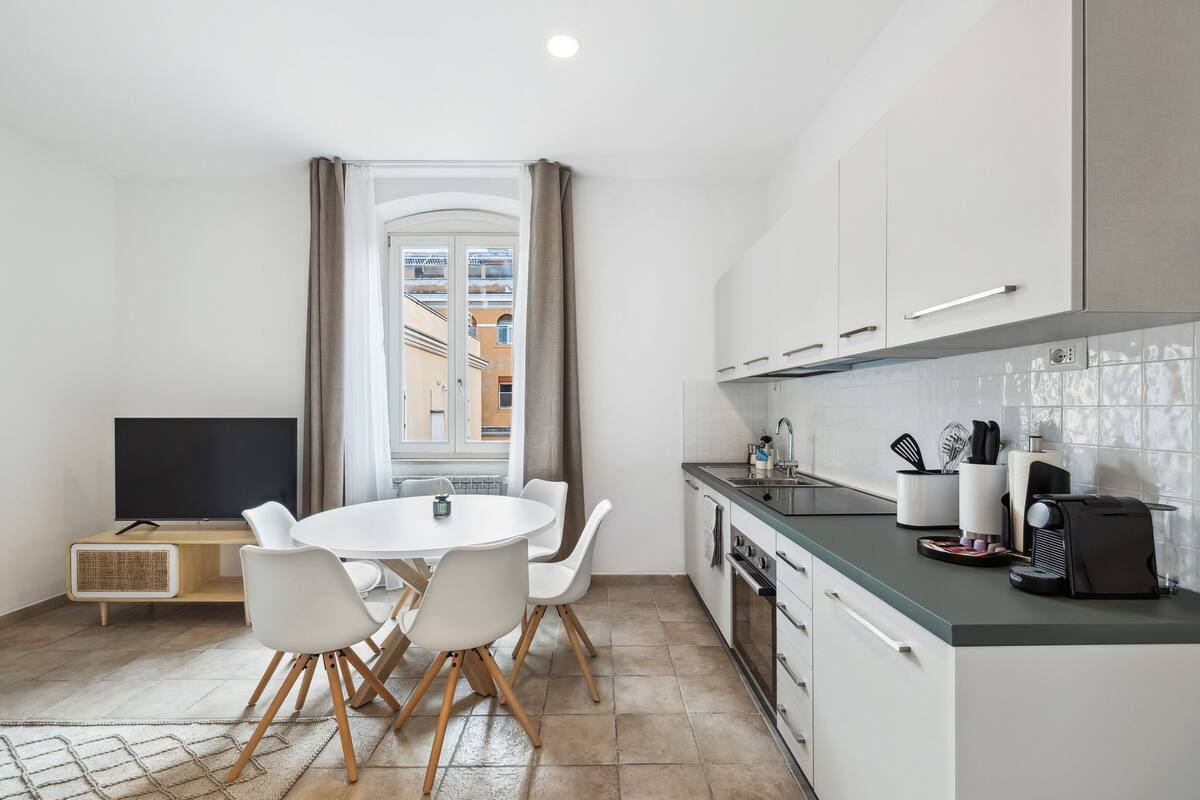 A modern kitchen with white cabinets, a dining table with chairs, and a large window with curtains.
