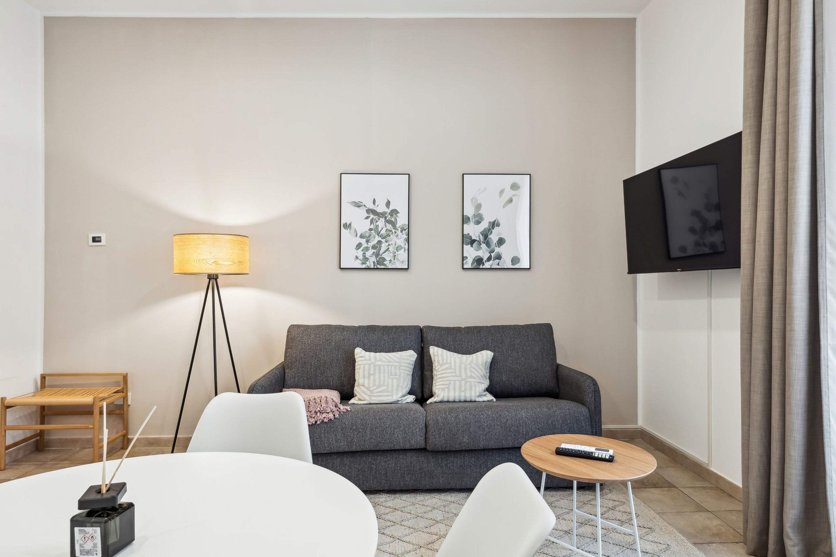 Un saló modern amb un sofà gris, butaques blanques, una taula de fusta petita i una làmpada de peu. Hi ha dues obres d'art emmarcades a la paret i un televisor muntat sobre el sofà.