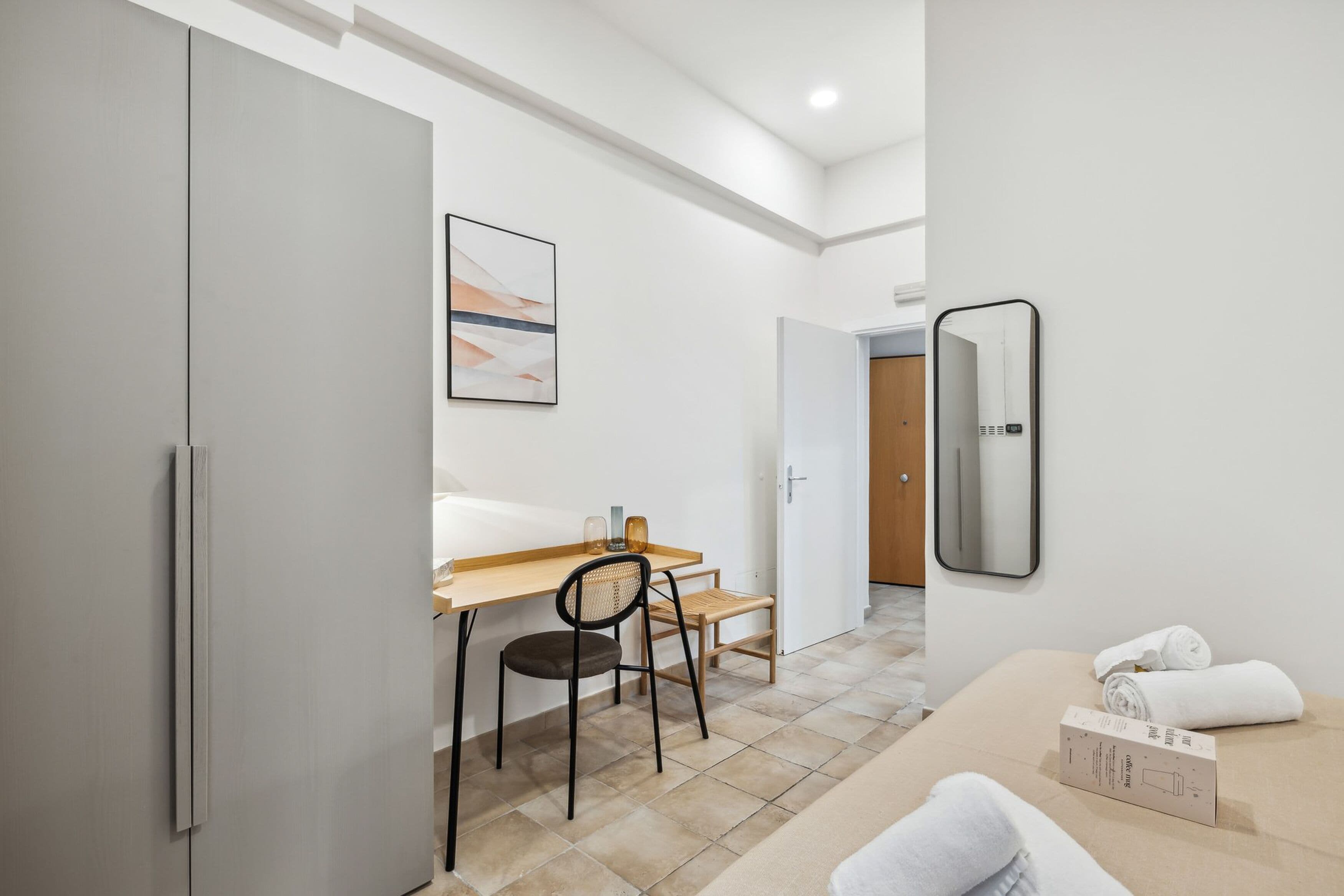 A modern hotel room with a bed, a desk, a chair, a mirror, and a painting on the wall.