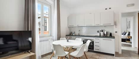 A modern kitchen with white cabinets, a dining area with a round table and chairs, and a TV on a wooden stand.