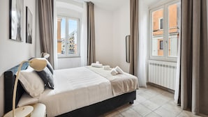 A hotel room with a bed, a bedside table, a lamp, a window with curtains, and a view of buildings outside.