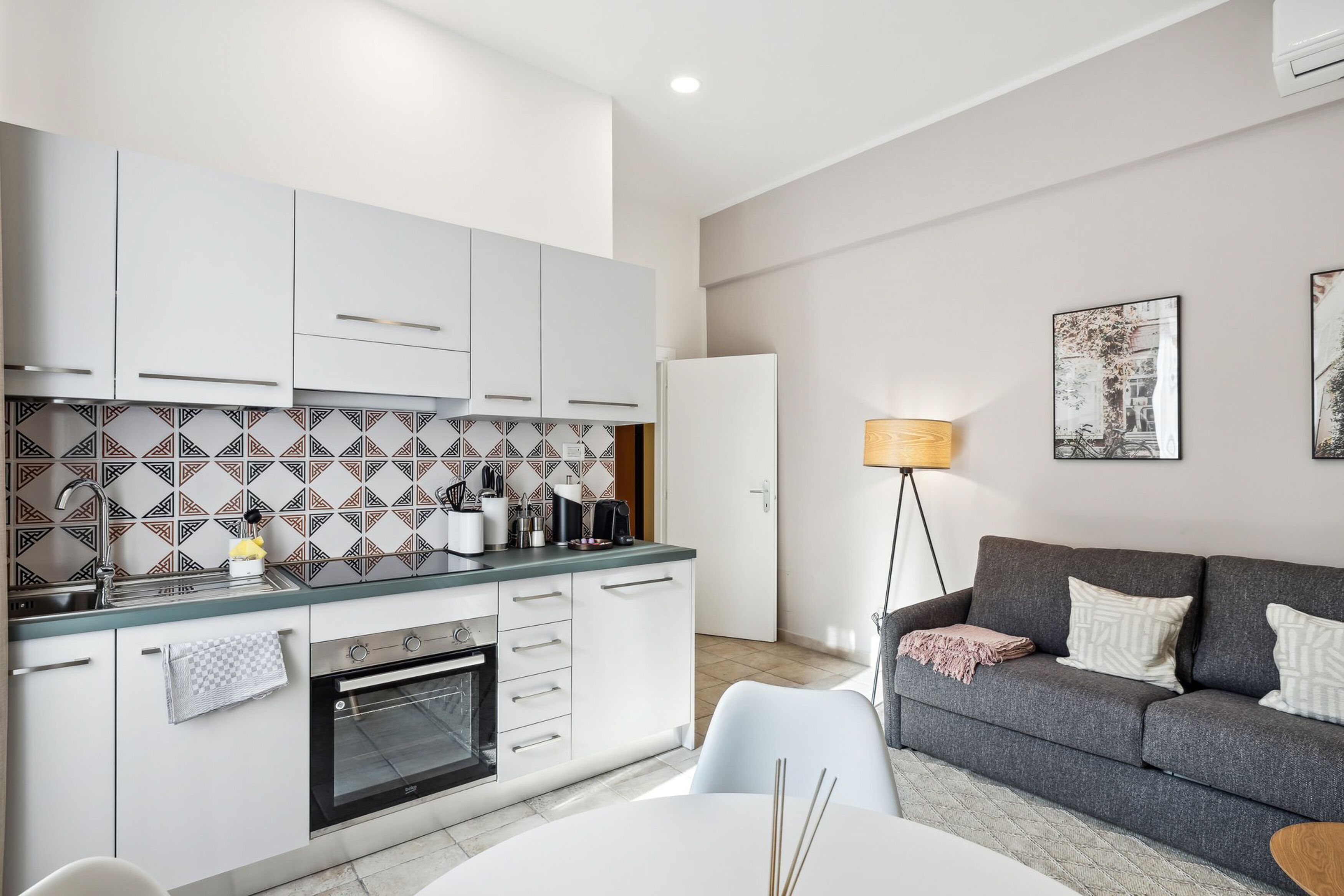Una cocina moderna con armarios blancos, horno empotrado y salpicadero de azulejos. Hay un sofá gris y una lámpara de pie en la zona de estar.