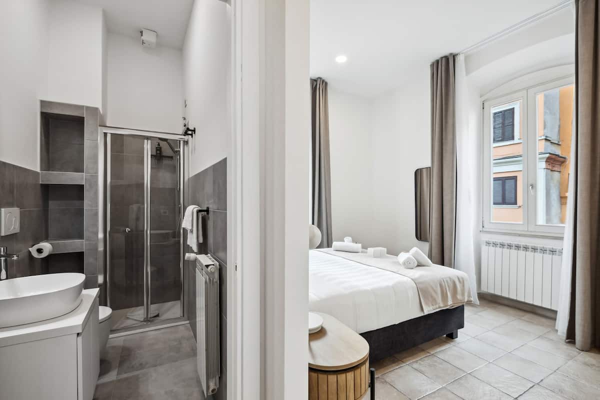 A modern bathroom with a shower, toilet, and sink. The room has a large window with curtains and a bed with white linens.