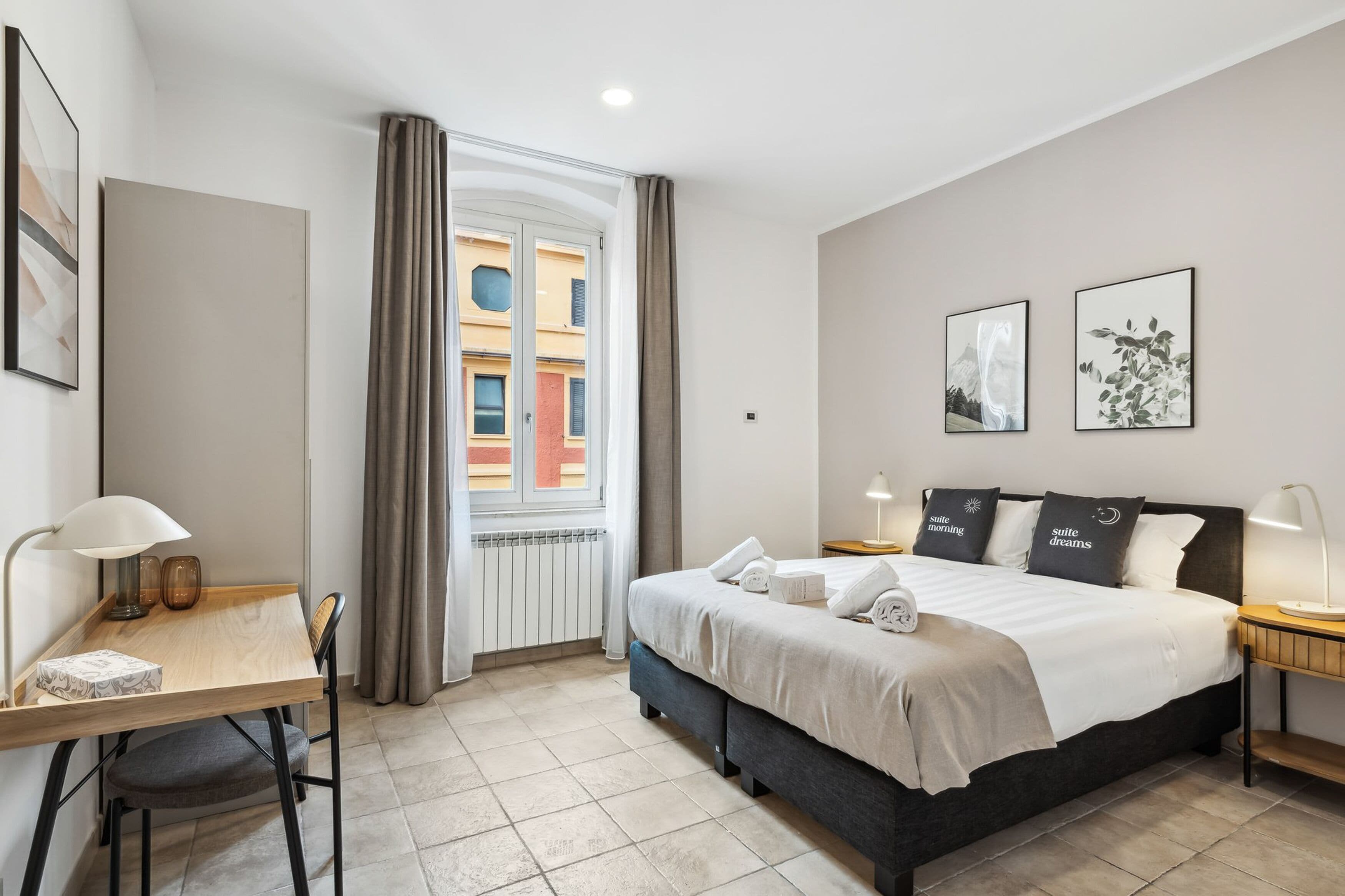 Una habitación de hotel con cama, escritorio, silla, ventana con cortinas y obras de arte en las paredes.