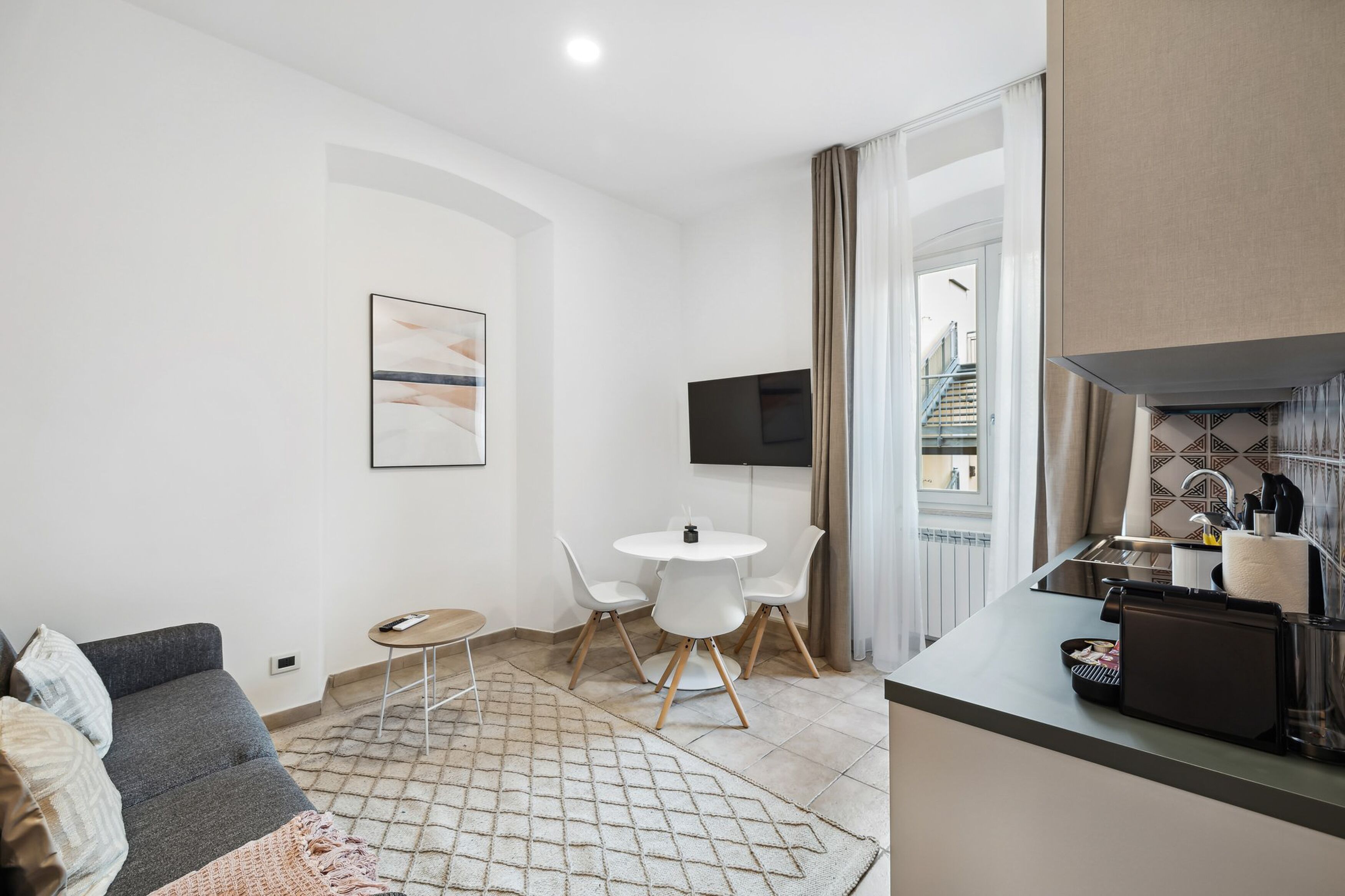 A modern living room with a sofa, a coffee table, and a TV. There is a dining area with chairs and a small table. The room has a kitchen area with a sink and a countertop.