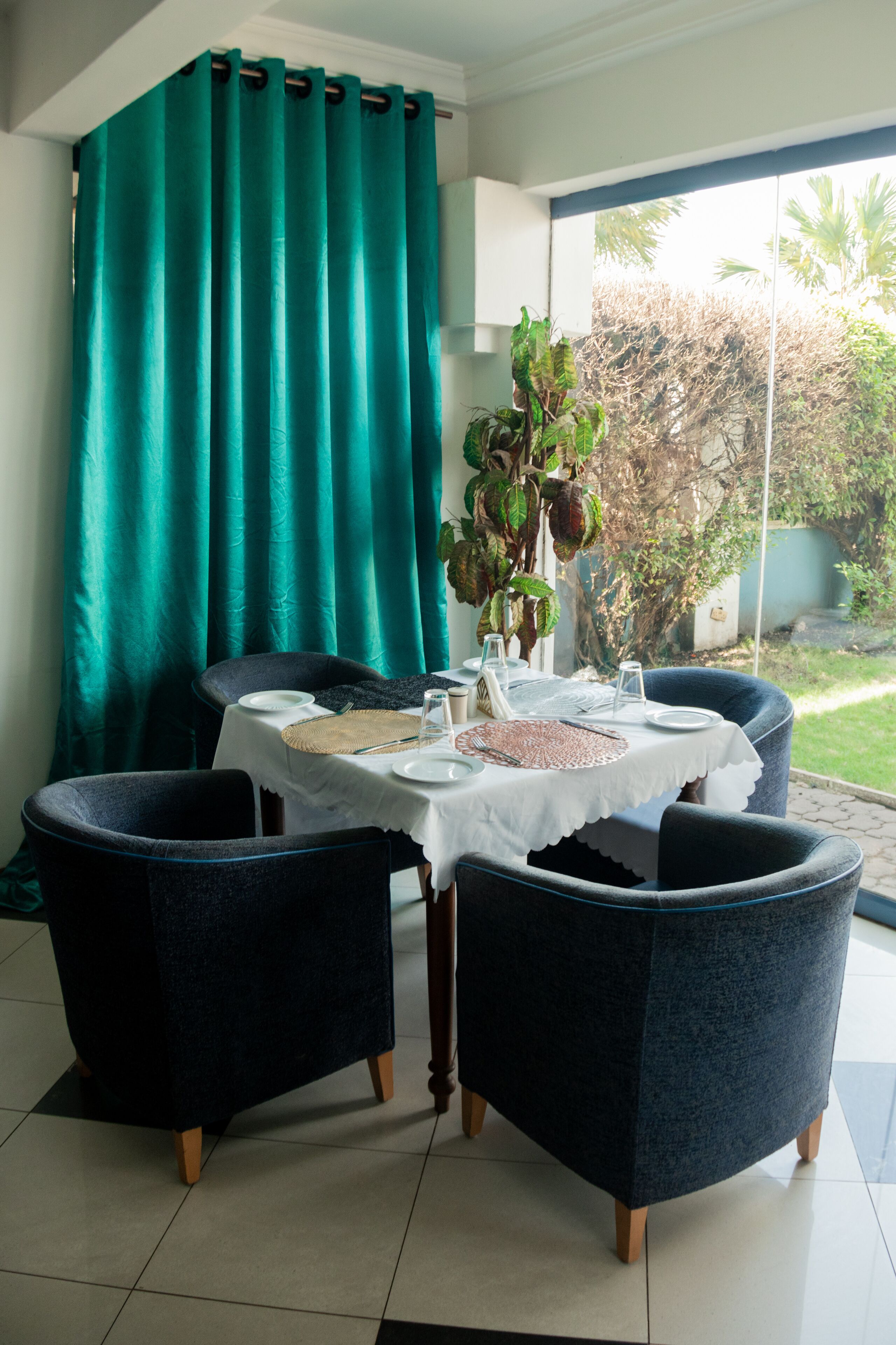 A dining area with a table set for four, surrounded by dark blue chairs, a teal curtain, and a view of greenery outside.