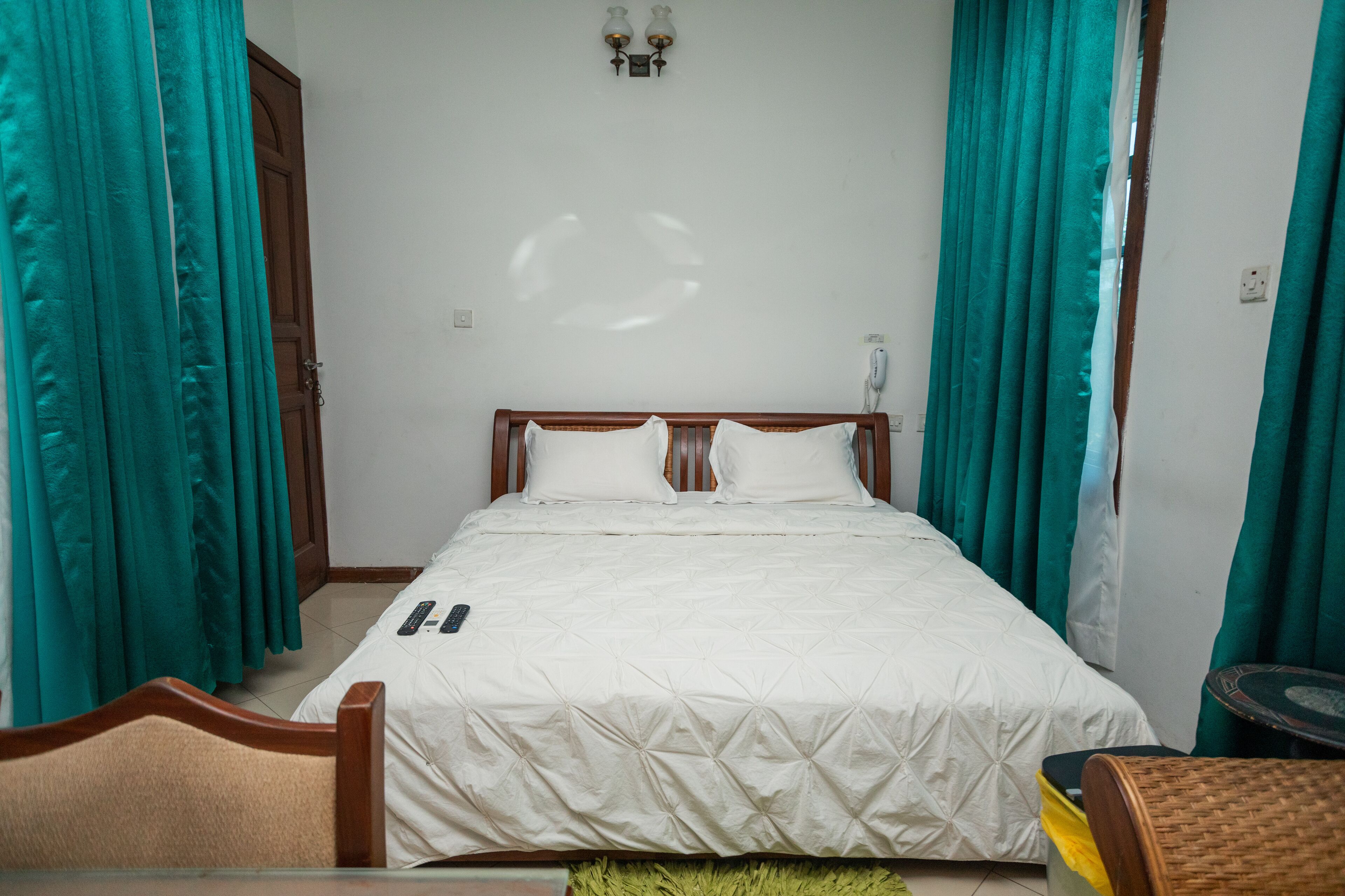 Ein ordentlich bezogenes Bett mit weißer Daunendecke, einem hölzernen Kopfteil und zwei Kissen. Auf dem Bett liegt eine Fernbedienung, und im Vordergrund steht ein Korbsessel.