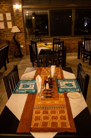 A dining table set for four with a patterned runner, chairs, and a lamp in a brick-walled room. - Sian Simba River Lodge (Victoria Falls)
