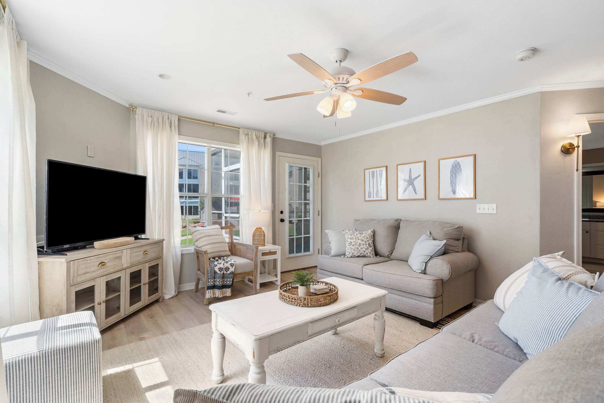 Ett vardagsrum med en platt-TV, en takfläkt, en soffa, ett soffbord och ett fönster med gardiner.