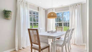 A dining area with a white table and chairs, large windows with white curtains, and a hanging wicker light fixture.