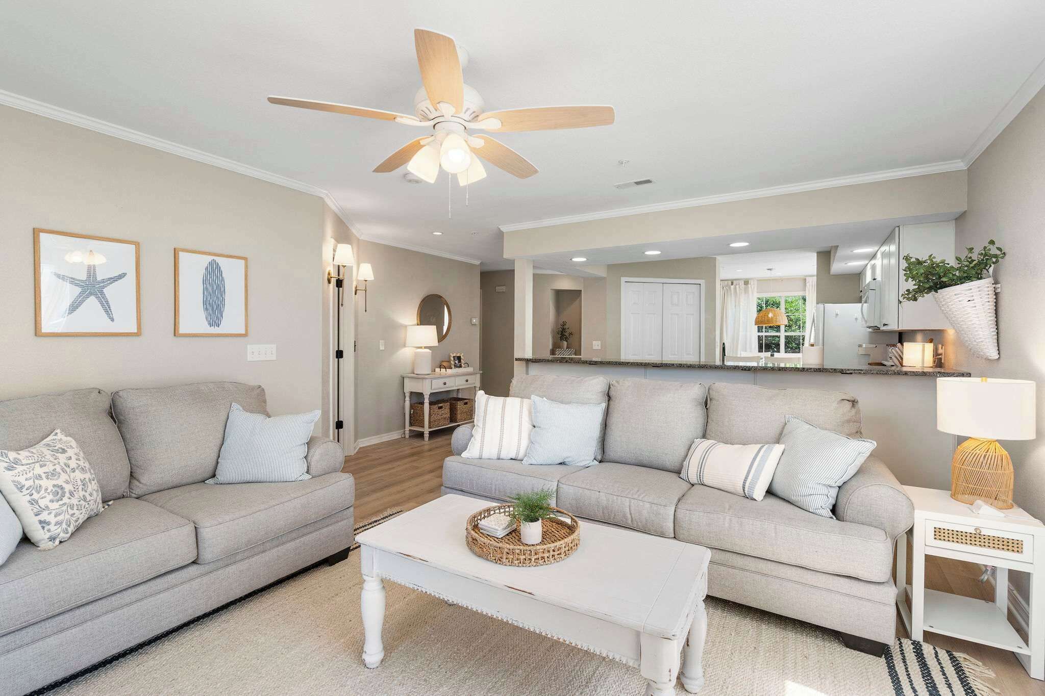 A living room with two sofas, a coffee table, and a ceiling fan. There is a kitchen visible in the background.