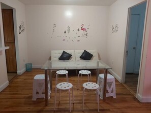 A dining area with a glass table, four stools, a sofa with two black cushions, and a wall with a flamingo mural.