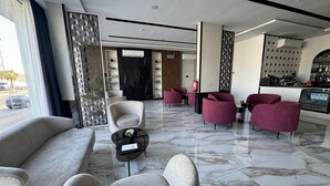 Un hall d’entrée moderne avec un sol en marbre, des canapés gris et des fauteuils rouges. On y trouve un comptoir d’accueil, et on aperçoit un coin cuisine à l’arrière-plan.