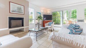 A living room with a fireplace, a television, a sofa, and a dining area with a view of greenery. - Madame Vacances Villas Club Royal 17 (Moliets-et-Maa)