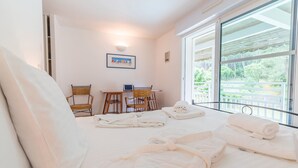 A hotel room with a bed, two wicker chairs, a small table, a laptop, and a view of greenery through large windows. - Madame Vacances Villas Club Royal 17 (Moliets-et-Maa)