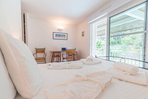 Une chambre d’hôtel avec un lit, deux fauteuils en osier, une petite table, un ordinateur portable et une vue sur la verdure grâce à de grandes fenêtres.
