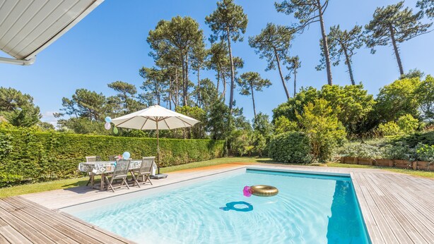 A rectangular swimming pool with a wooden deck, surrounded by lush greenery and tall trees. - Madame Vacances Villas Club Royal 17 (Moliets-et-Maa)