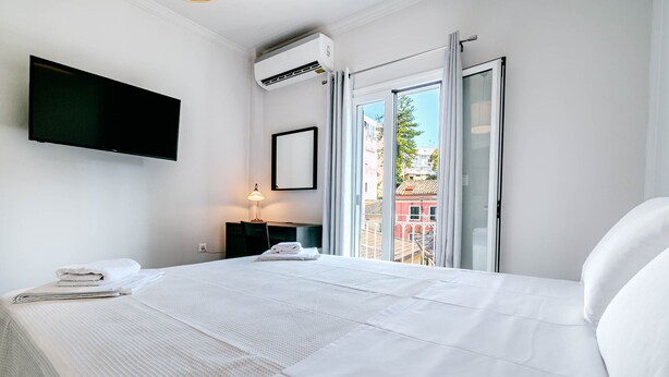 A hotel room with a large bed, a flat-screen TV mounted on the wall, an air conditioning unit, a bedside table with a lamp, and a window with a view of buildings outside. - Alex Home by Corfuescapes (Corfu)