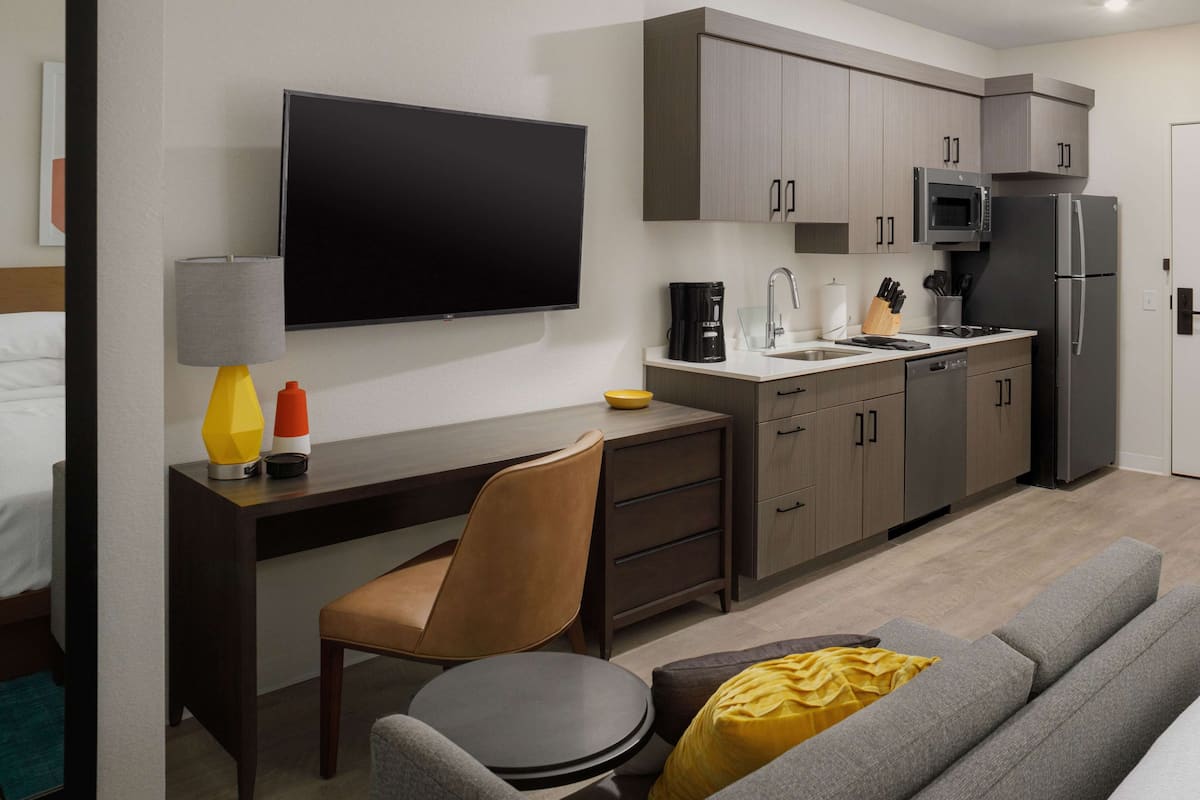 Une chambre d’hôtel moderne équipée d’une télévision à écran plat, d’un bureau avec une chaise, d’un canapé, d’un coin cuisine avec un four à micro-ondes et un réfrigérateur, et d’un lit.