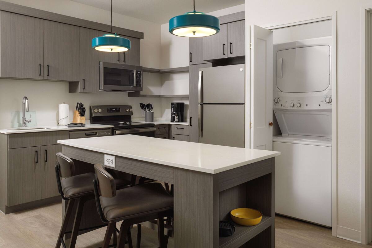 Une cuisine moderne avec des meubles gris, un îlot central doté de tabourets, un four encastré, un micro-ondes et un réfrigérateur en acier inoxydable.