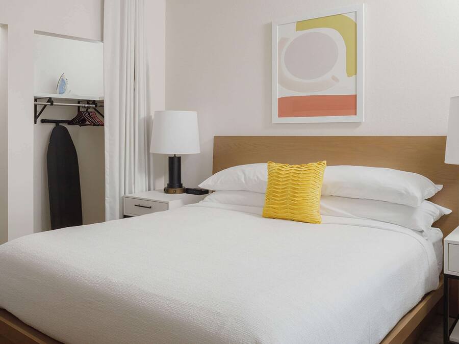 A neatly made bed with white bedding and a yellow pillow, a bedside lamp, a wall-mounted abstract artwork, and a clothing rack with hanging garments.