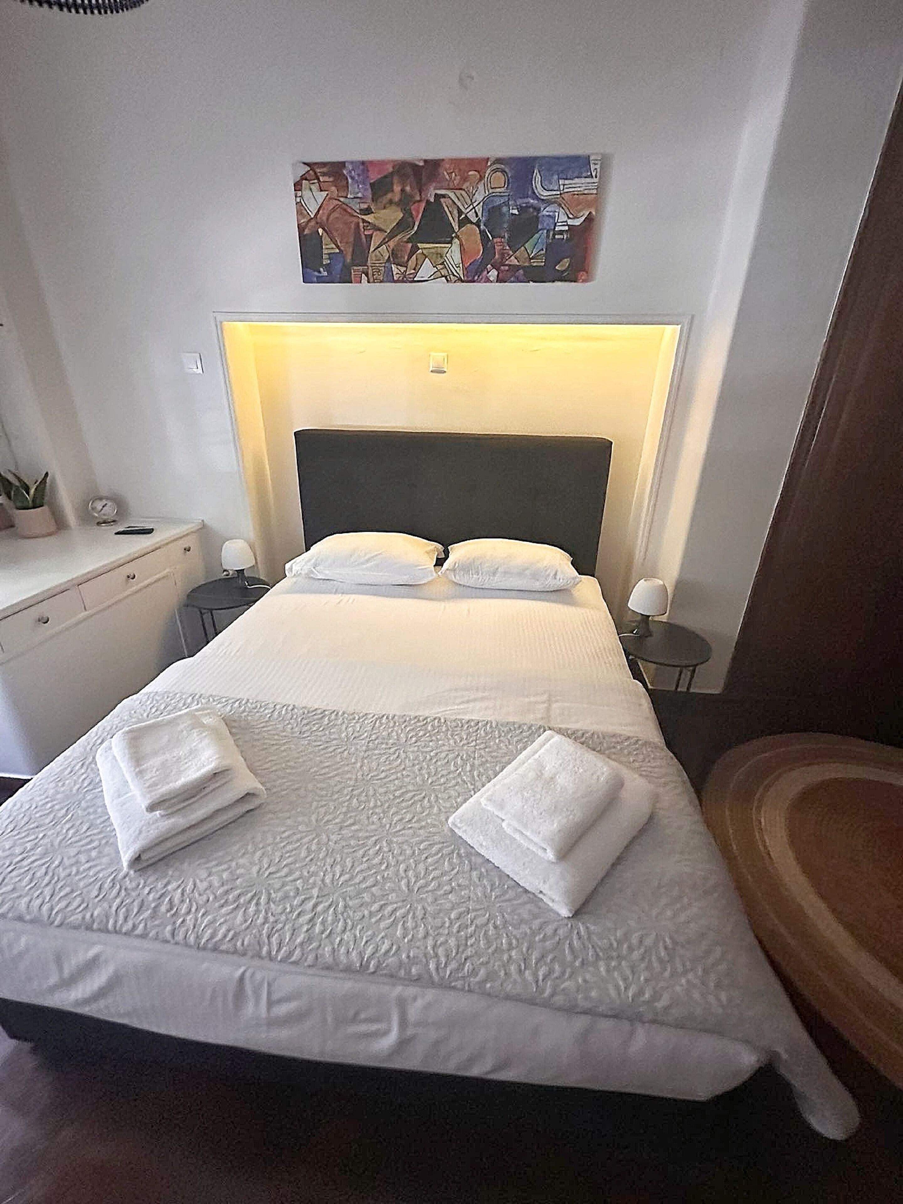 Una cama bien hecha con sábanas blancas y toallas dobladas, una mesita de noche con una lámpara, y un cuadro abstracto colgado en la pared sobre la cama.