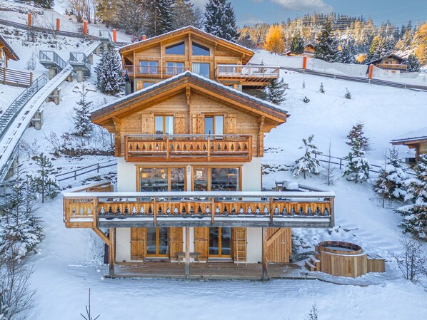 A two-story wooden chalet with a balcony, a hot tub, and a ski lift in the background. - Chalet Sapin Argenté (Riddes)