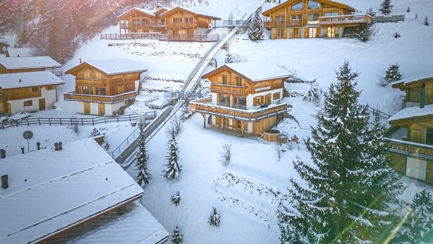 En snötäckt semesteranläggning med stugor byggda av trä, en skidlift och en imponerande bergskuliss.