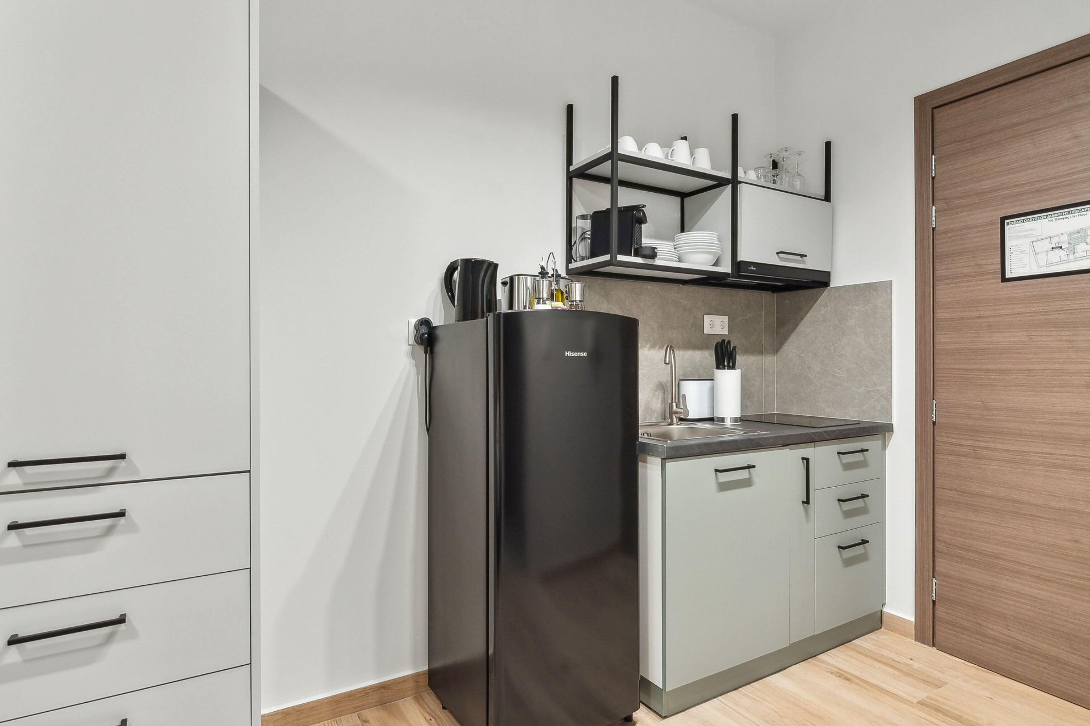 Cocina compacta con un refrigerador negro, horno empotrado y una pequeña encimera. Hay armarios con manijas negras y una puerta de madera.