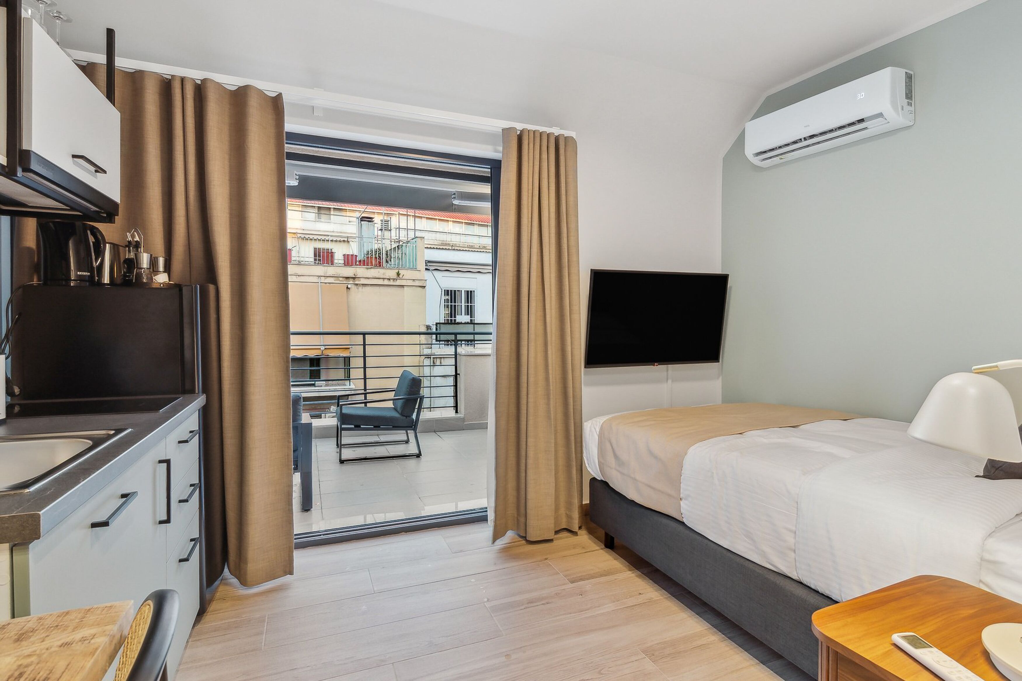 Une chambre d’hôtel comprenant un lit, une télévision, un appareil de climatisation, un coin cuisine et un balcon avec une chaise et une table.