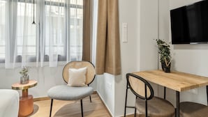 A modern living room with a sofa, a wooden dining table, a TV mounted on the wall, and a window with sheer curtains. - Limehome Athens Solonos City Center (Athens)