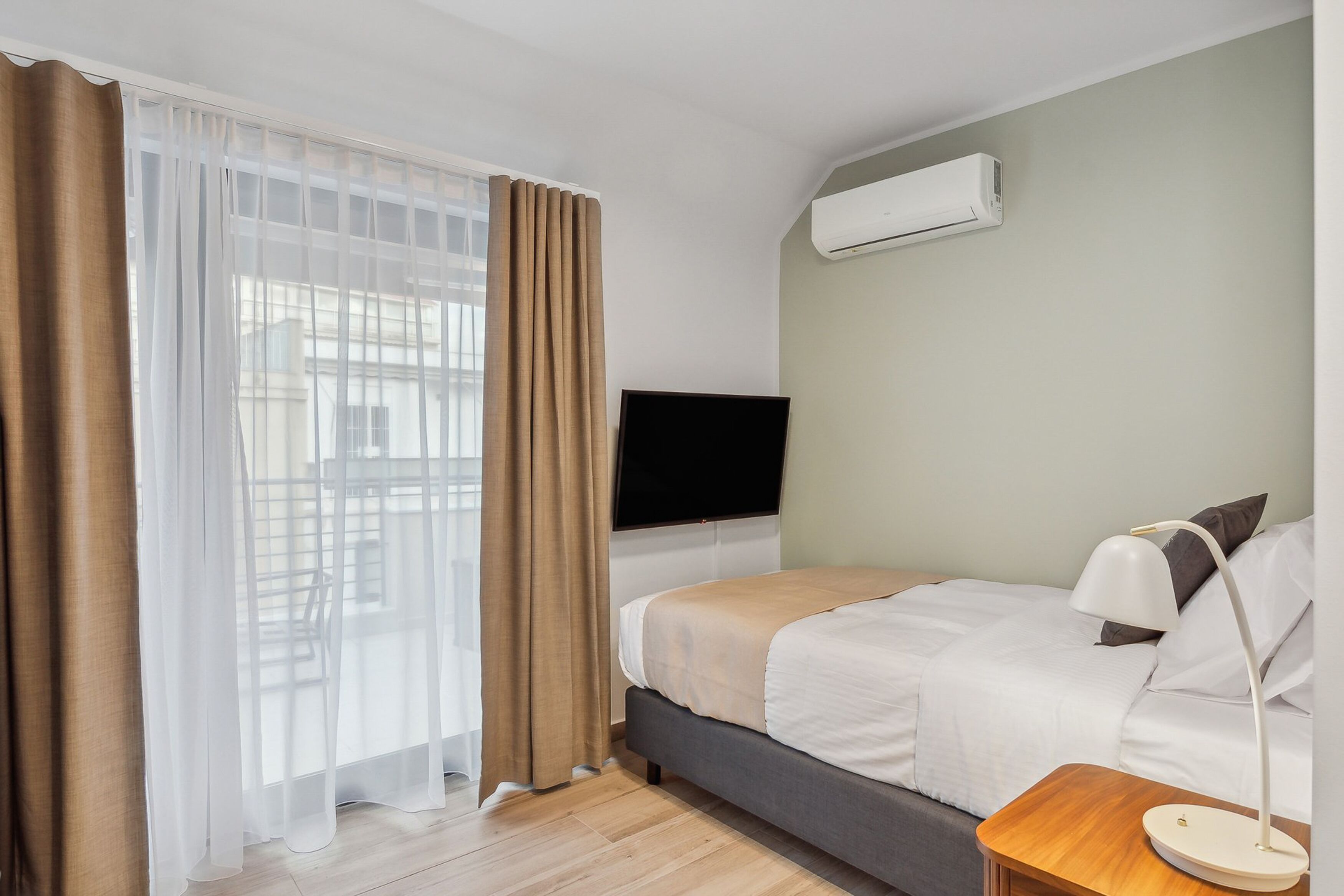 A hotel room with a bed, a TV mounted on the wall, an air conditioning unit, a wooden nightstand, and a window with sheer curtains.