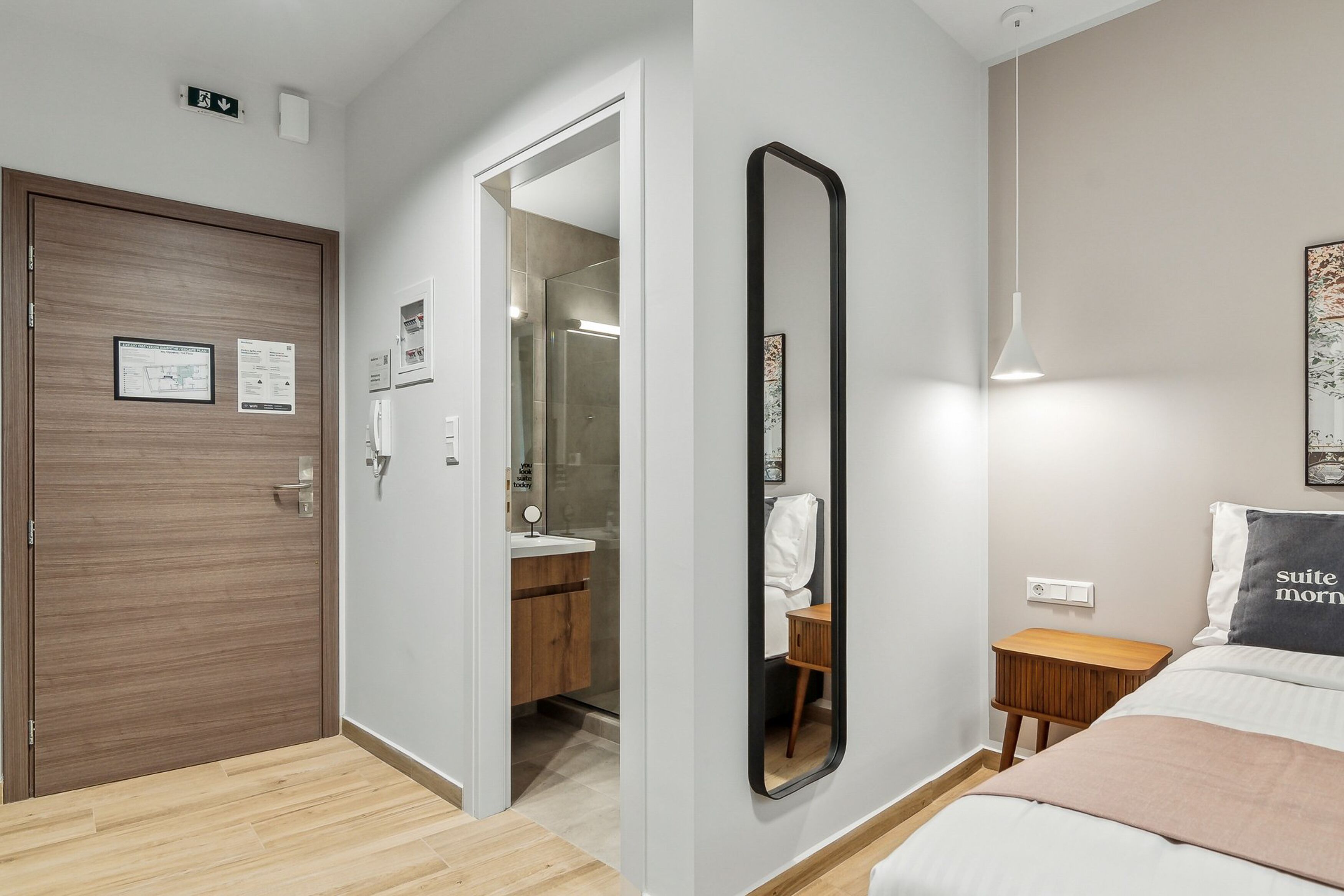 Habitación de hotel con puerta de madera, un espejo, una mesita de noche y una cama con almohada.
