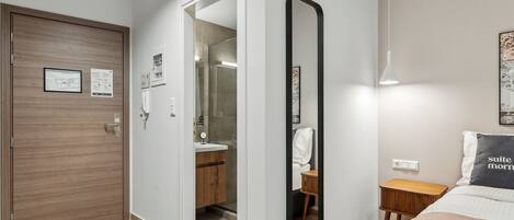 Habitación de hotel con puerta de madera, un espejo, una mesita de noche y una cama con almohada.